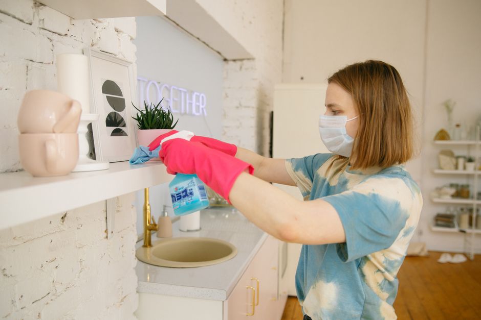 Woman wearing mask and gloves sanitizing shelves in a bright kitchen.